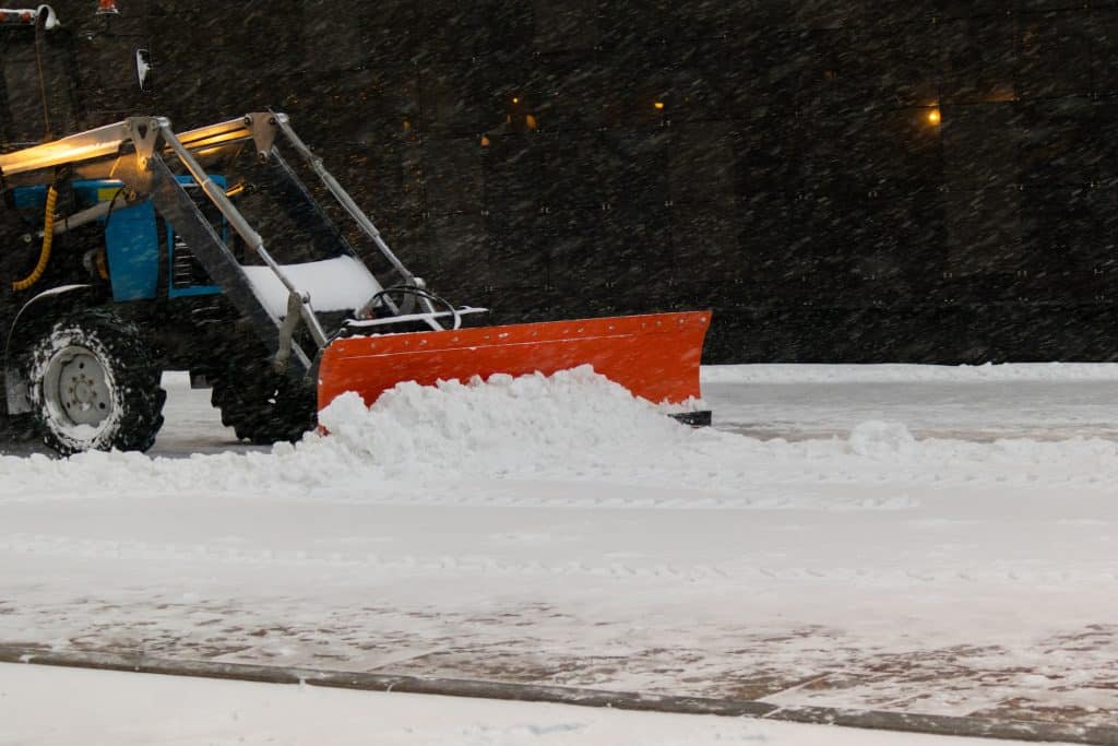 A front-end loader fitted with a bright orange plow blade pushes fresh snow across a wet pavement under heavy snowfall, set against a dark urban building backdrop.